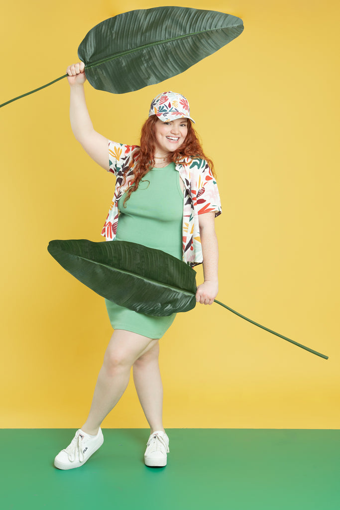 A red haired woman holds two palm leaves while wearing a coordinated ensemble of in a bright and bold summer pattern