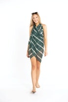 Young blonde woman is smiling wearing a colorful, striped sarong wrapped around her neck as a coverup dress. She has sunglasses propped on top of her head and is wearing strappy sandals.