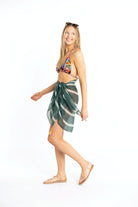 Tall, blonde, young woman is smiling as she is strutting in a teal striped sarong coverup and a multi-colored bikini top. She has sunglasses on her head and is wearing strappy sandals on her feet.