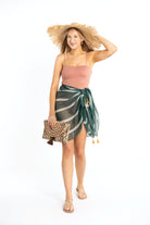 Tall white girl with blonde hair wearing a swimsuit and colorful, striped sarong coverup. Her hand is holding on to the brim of the braided woven raffia hat on her head. Her other hand is holding a colorful clutch with tassel trim.