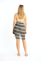 Tall, blonde, young woman is smiling as she is posing from behind. She is wearing a teal striped sarong coverup and a multi-colored bikini top. She has sunglasses on her head and is wearing strappy sandals on her feet.