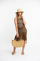 Young blonde model wears a brown sheer beach coverup and a woven raffia straw bucket hat holds a braided cane straw tote bag with circular wooden handle.