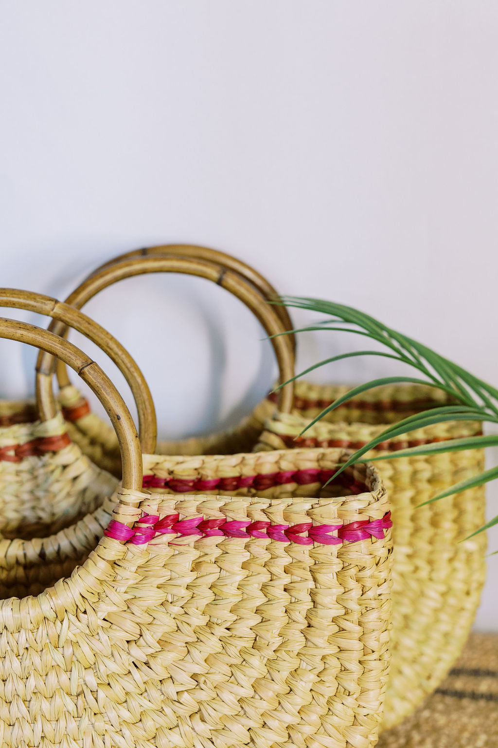 Closeup of two hand-crafted cane tote bags with circular bamboo polished handles. Front tote has a pink stripe near the top opening and the background tote has a rust stripe accent.